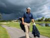 A Florida golfer carrying a waterproof rain jacket and golf bag on a sunny course with dark storm clouds approaching