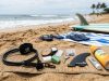 A complete surfboard accessories kit laid out on the beach including a leash, wax, fin key, and sunscreen