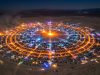 Aerial view of Black Rock City's circular street grid glowing with lights at night during Burning Man 2026 in the Nevada desert